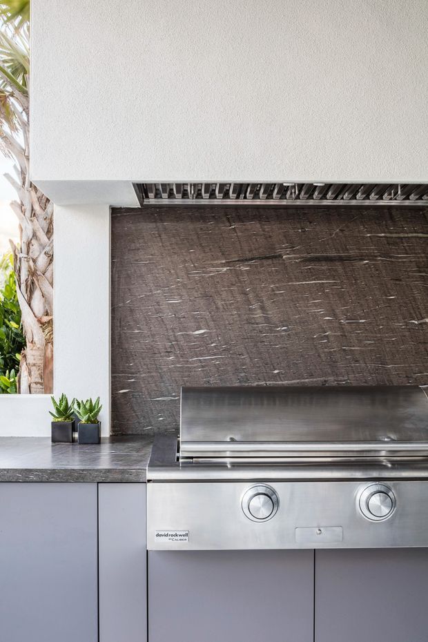 Close-up of a contemporary outdoor cooking area featuring a built-in stainless-steel barbecue with two control knobs, dark textured stone or tile splashback, grey cabinetry, and a grey benchtop. White rendered overhead structure and tropical planting are visible at the edge of the image.