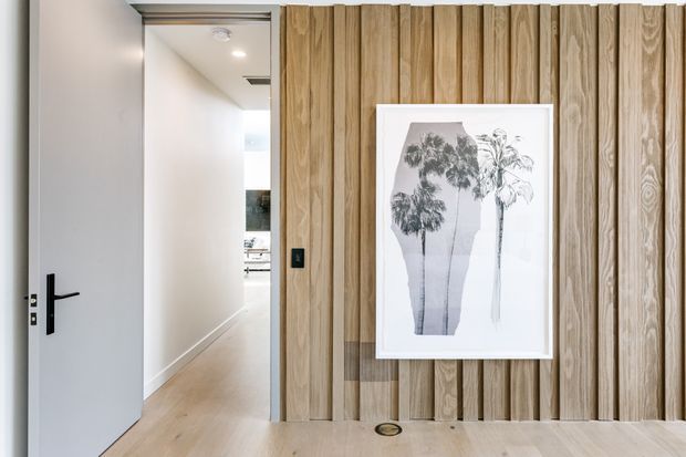 A bright contemporary interior hallway or entry passage with pale timber flooring and a striking vertical timber feature wall. The wall is clad in narrow, varied-grain timber boards and displays a framed monochrome botanical artwork, with a recessed floor light at its base. A simple white door with black hardware opens toward a well-lit corridor and kitchen or living area beyond.