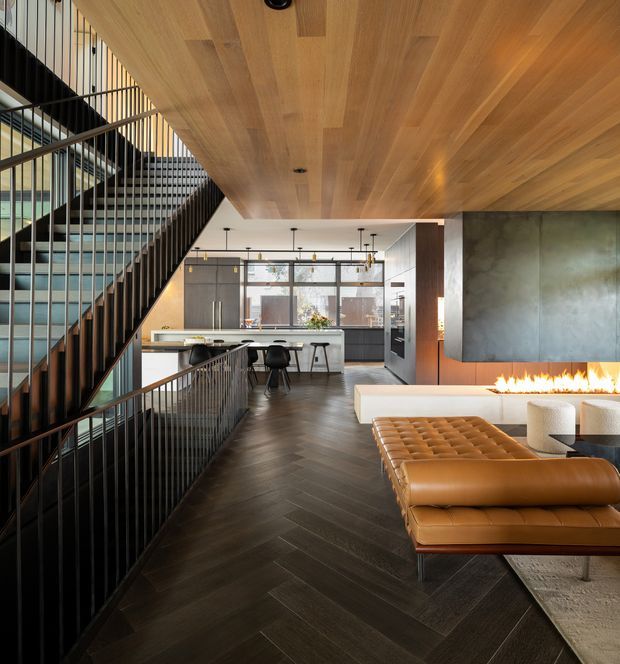 A refined open-plan contemporary interior viewed from a split-level living area, featuring a dramatic timber-lined ceiling and dark herringbone timber flooring. A black metal stair and balustrade frame the left side, leading toward a compact kitchen and dining zone with dark cabinetry, a pale island bench, and pendant lighting. The living area includes a caramel leather tufted ottoman and sofa, a pale rug, cylindrical stools, and a long linear fireplace set beneath a large grey concrete or stone feature wall. Strong glazing at the rear brings in natural light, while dark metal and timber details create a warm, sophisticated atmosphere.