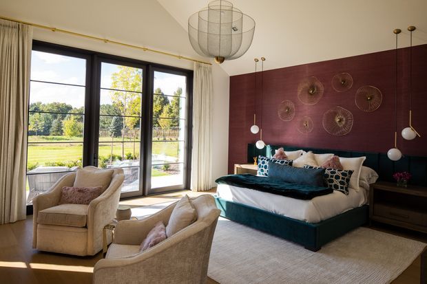 A refined bedroom with a large upholstered bed positioned against a deep burgundy feature wall decorated with sculptural circular wall pieces. Floor-to-ceiling black-framed glazed doors open to a green outdoor landscape, while cream curtains, plush armchairs, layered bedding, and a large neutral rug create a warm, luxurious sitting area within the room. Contemporary pendant lighting and dark bedside joinery complete the space.