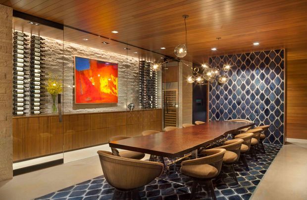 A richly detailed dining and wine room with a long sculptural timber dining table surrounded by woven armchairs. The room features extensive built-in wine storage along a textured stone wall, warm timber wall and ceiling panelling, mirrored panels, sculptural pendant lighting, colourful artwork, and a bold blue patterned feature wall and carpet.