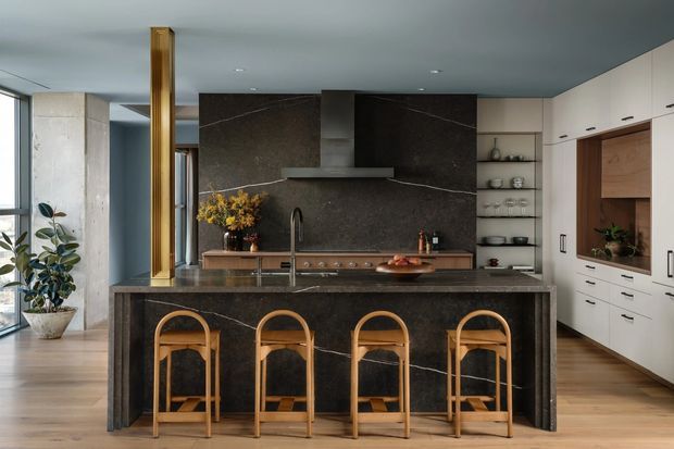 A sophisticated contemporary kitchen with a large dark stone island, integrated cooktop and sink, tall white cabinetry, open shelving, and a dark stone splashback with a sculptural rangehood. Four timber-and-cane bar stools line the island, while warm timber flooring and a vertical timber feature screen add contrast. Glazing and indoor plants bring natural light and greenery into the space.
