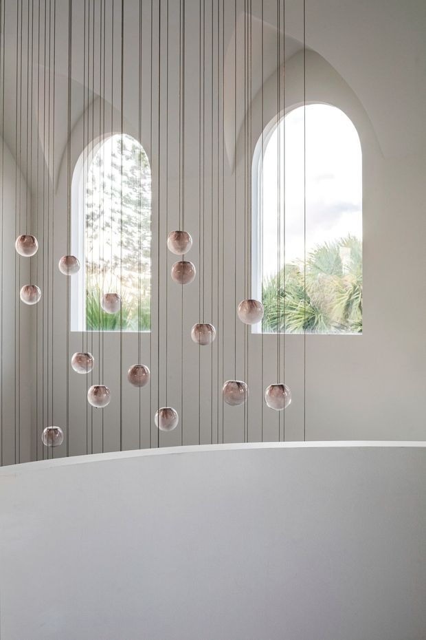 A serene, minimalist interior featuring a curved white architectural partition or balustrade in the foreground, a tall vertically panelled wall, and two large arched windows. Numerous small translucent spherical pendant lights hang at varied heights from long fine cords, creating a sculptural installation. Soft daylight, pale neutral finishes, and glimpses of greenery through the windows give the space an airy, refined atmosphere.