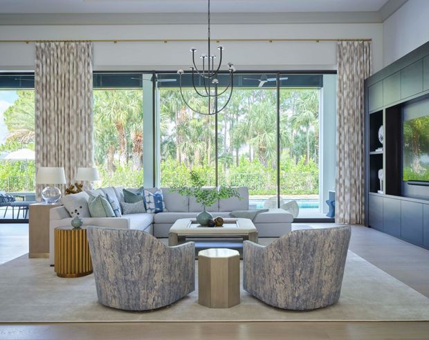 Bright, spacious contemporary living room with floor-to-ceiling glazing opening onto a lush outdoor setting and pool. The room features a large pale sectional sofa, sculptural upholstered lounge chairs, patterned full-height curtains, a dark built-in media wall, a contemporary chandelier, and neutral stone-toned finishes.