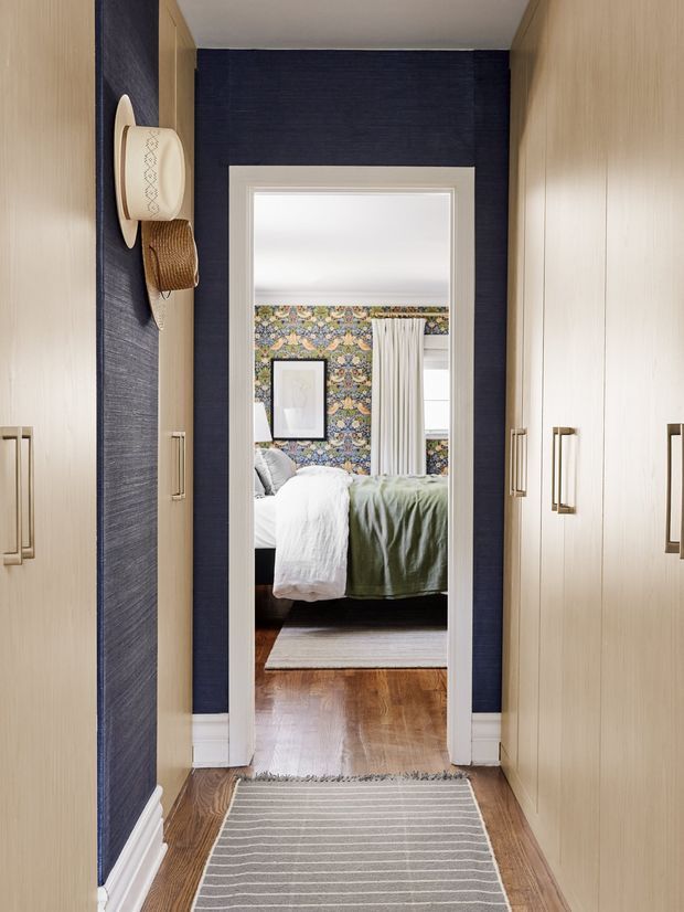 A narrow, refined walk-in wardrobe or dressing passage with full-height light timber cabinetry on both sides, brass-toned handles, dark navy textured wall covering, white architectural trims, and timber flooring. A striped runner leads through a doorway into a bedroom, where a patterned feature wall, curtains, framed artwork, and a bed with white and sage-green bedding are visible.