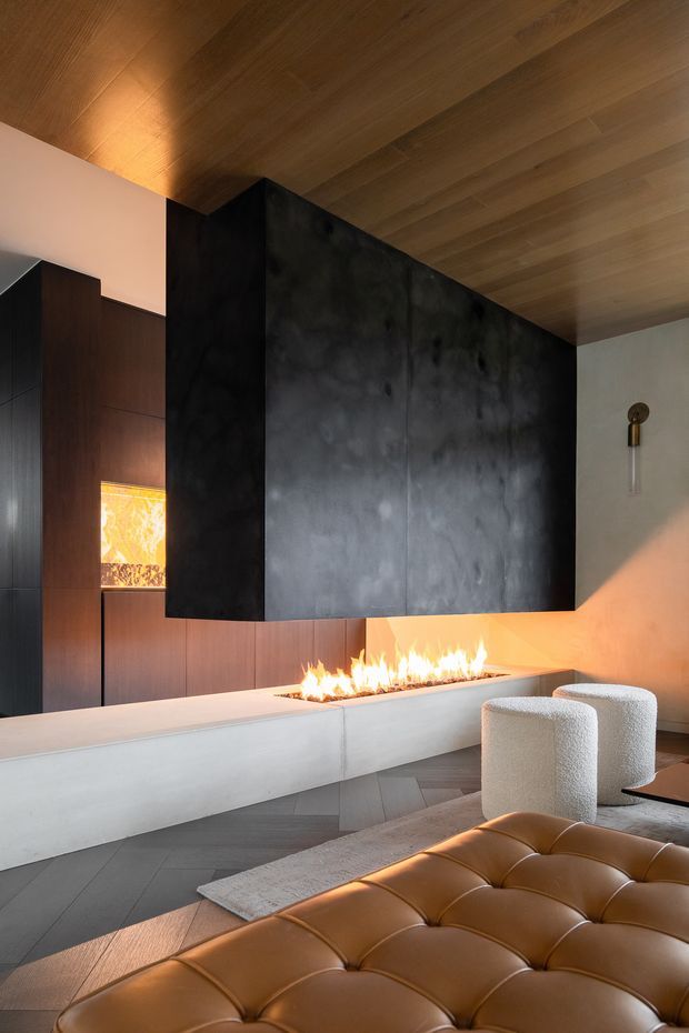 A refined contemporary living room centred on a long linear fireplace set into a pale stone or concrete hearth. A dramatic black monolithic chimney hood projects from the wall beneath a warm timber-lined ceiling. Dark timber wall panelling, pale walls, grey geometric flooring, boucle lounge chairs, and a tan leather tufted ottoman create a luxurious, moody interior with warm firelight.