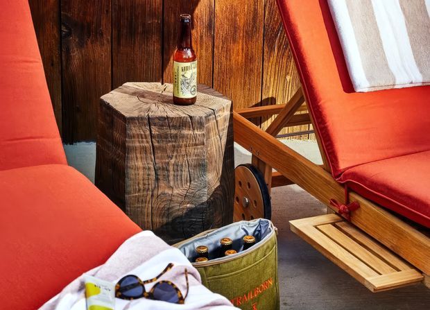 A close-up outdoor seating arrangement with two red-cushioned wooden lounge chairs around a rustic tree-trunk side table. A beer bottle, sunglasses, folded towel, and a small barrel-style cooler add a relaxed recreational atmosphere. Dark vertical timber paneling forms the background, with warm natural wood tones throughout.