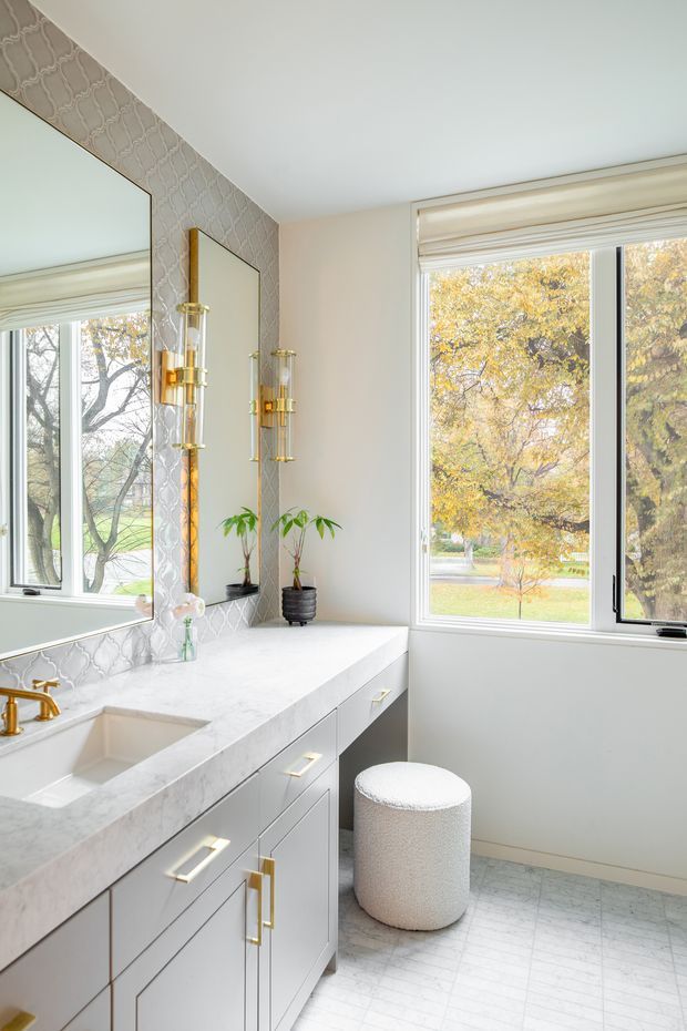 Bright, refined bathroom vanity area with a long white stone benchtop, integrated sink, pale grey built-in cabinetry, brass fixtures and hardware, patterned light-grey wall tile, and a matching pale tiled floor. Large windows with roller blinds bring in abundant natural light and overlook autumn trees. A large wall mirror, decorative brass sconces, potted plants, and a round upholstered stool complete the space.