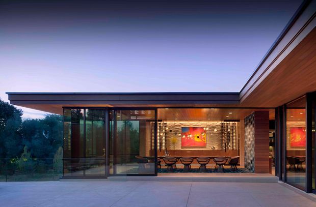 Contemporary low-profile residence viewed from a broad paved terrace at dusk. The open glazed facade reveals a warm dining area with a long table, sculptural chairs, timber wall cladding, exposed timber ceiling elements, patterned masonry feature walls, and artwork. The building has strong horizontal rooflines, extensive floor-to-ceiling glazing, dark slender frames, and an indoor-outdoor connection.