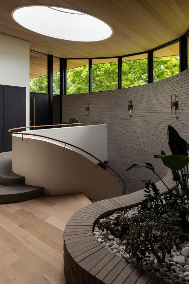 Interior view of a refined modern stairway and landing. A sweeping curved staircase with a white plaster balustrade and slim timber handrail descends around a large circular indoor planter filled with greenery and white stones. The space features warm timber ceiling and flooring, dark metal-framed clerestory glazing, a pale grey brick feature wall with integrated wall lights, and a circular roof skylight.