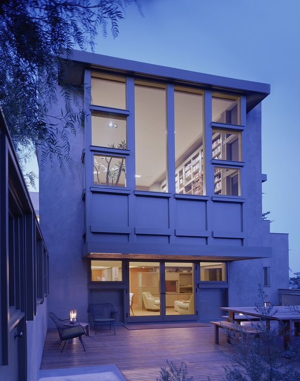 A twilight view of a narrow multi-storey contemporary home with a deep blue painted facade, large vertically proportioned windows, and a projecting upper-level volume. The ground floor opens onto a timber deck courtyard with outdoor seating and dining furniture. Warm interior lighting reveals a living area and extensive built-in shelving through the glazing, while mature foliage frames the building.