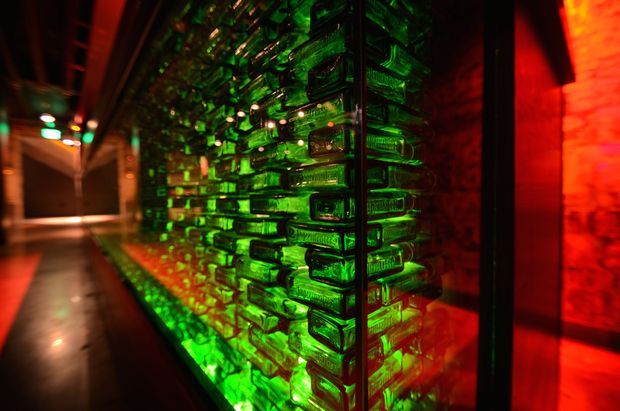 Dramatic close-up of a translucent textured glass block wall or partition in a dark corridor, illuminated with vivid green and red accent lighting. The glass blocks form a sculptural architectural feature, with a reflective dark floor and exposed brick surfaces visible around the passage.