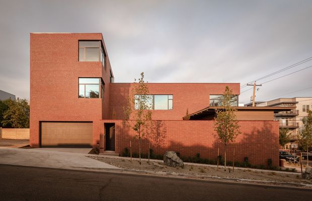 Street-facing exterior view of a contemporary multi-level brick residence. The building has a strong rectilinear composition, a tall projecting volume, broad horizontal wings, large modern windows, an integrated garage, and layered roof or terrace elements. A brick boundary wall, young trees, paved driveway, utility lines, and neighbouring urban buildings are visible.