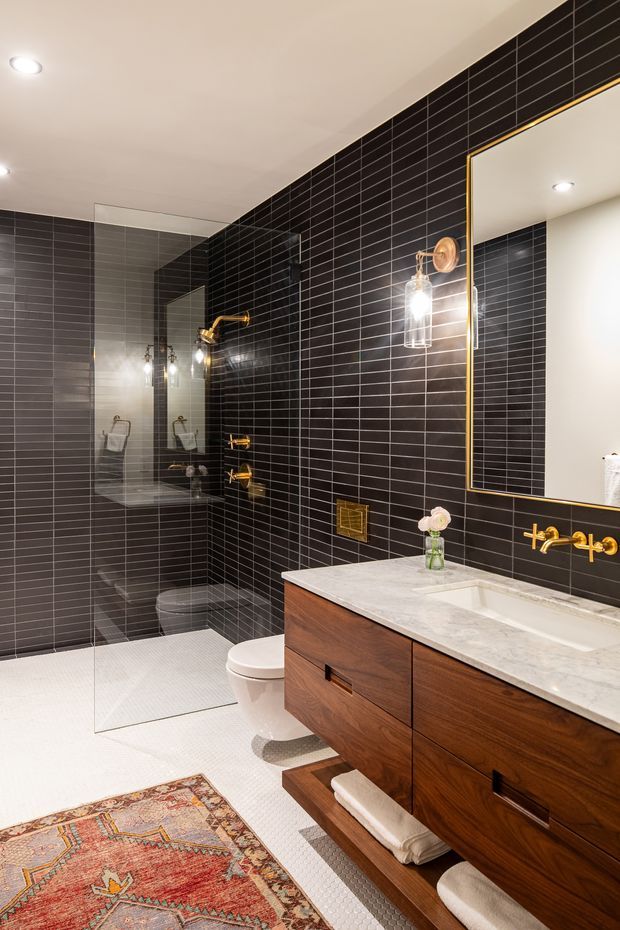A stylish modern bathroom with black horizontal rectangular wall tiles, a white small-format tiled floor, and a frameless glass walk-in shower. A floating dark timber vanity with a pale stone benchtop, integrated white basin, brass wall-mounted taps, brass-framed mirror, and matching brass lighting creates a warm, refined contrast. A patterned rug and folded towels add softness and colour.