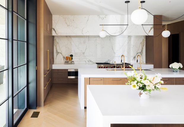 A refined contemporary kitchen with a large white island, warm timber cabinetry, and dramatic veined marble-style wall surfaces. Black-framed glazed doors and windows line the left side, while sculptural globe pendant lights hang above the island. Brass-toned tapware and hardware add a warm accent, and the timber floor is laid in a herringbone pattern.