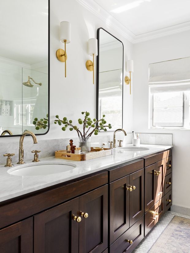 Bright, refined bathroom with a long double vanity featuring dark wood cabinetry, a white marble-look benchtop, integrated basins, brass faucets and hardware, and paired tall rounded-rectangle mirrors. White walls with decorative ceiling moulding, globe-like wall sconces, a window with a light blind, and a glass shower enclosure are visible. The styling is classic and elegant with warm brass accents and natural greenery.