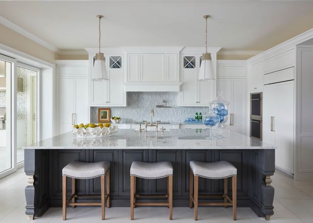 Bright, spacious traditional kitchen featuring a large dark-painted island with three upholstered timber bar stools, white raised-panel cabinetry, a pale herringbone tile backsplash, and a light stone countertop. Two pendant lights hang above the island, with integrated appliances and full-height storage along the walls. Glazed doors and patterned adjacent space are visible at left.