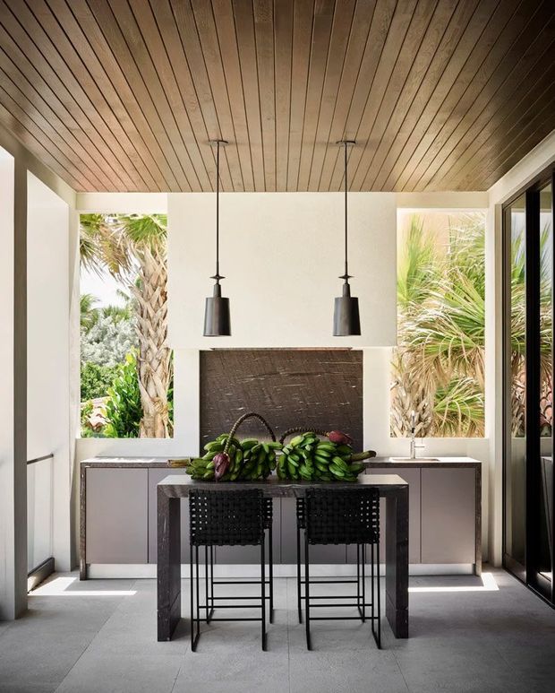 A bright, compact kitchen or entertaining space with a central dark stone island and two black woven bar stools. The room features white walls and cabinetry, a full-height dark textured backsplash, large glazed openings to lush tropical planting, slim black pendant lights, and a warm timber-lined ceiling. Pale large-format flooring and strong indoor-outdoor connections create a refined, tropical-modern atmosphere.