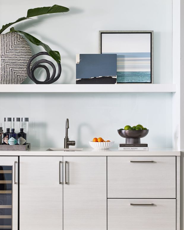 A bright, refined built-in kitchen or bar cabinetry vignette with pale wood-grain drawers and doors, white benchtops and open shelving, a compact stainless-steel tap, and a wine or beverage refrigerator at left. The shelves display bottles, bowls of fruit, books, framed artwork, sculptural objects, and a large leafy plant. The overall composition is clean and contemporary with a soft neutral palette and subtle coastal artwork.