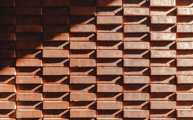 Close-up architectural detail of a textured red-brown brick wall. The bricks are arranged in a projecting, staggered pattern that creates deep horizontal and diagonal shadow lines, producing a strongly tactile geometric facade.