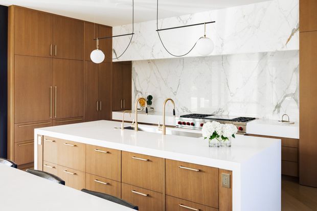 Bright, refined contemporary kitchen with extensive warm timber cabinetry, a large white island with integrated sink and brass-toned tapware, and a dramatic white marble-look splashback and rangehood surround. Minimal linear pendant lighting and polished surfaces create a clean, luxurious feel.