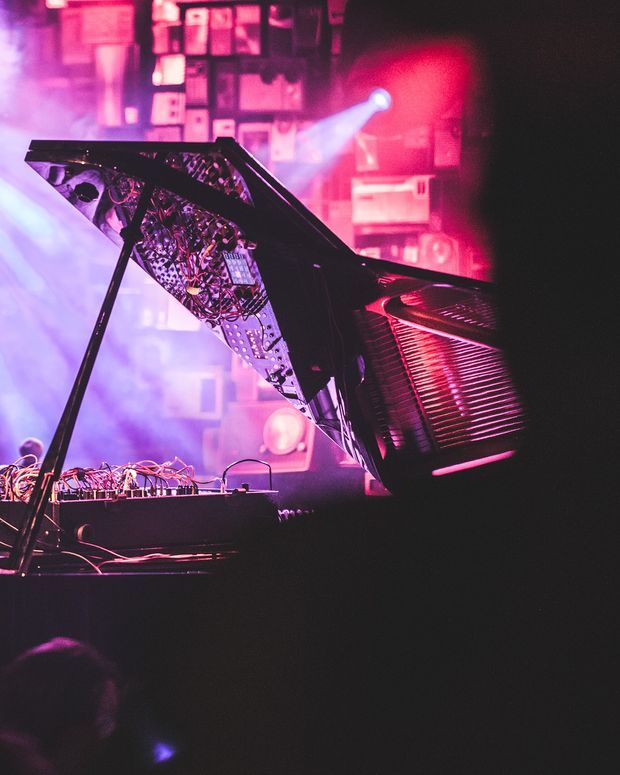 A dark, dramatic concert or performance setting featuring an open grand piano with its internal strings and mechanical components exposed. Pink, red, purple, and blue stage lighting, haze, audio equipment, cables, and a silhouetted audience create a vivid atmospheric scene.