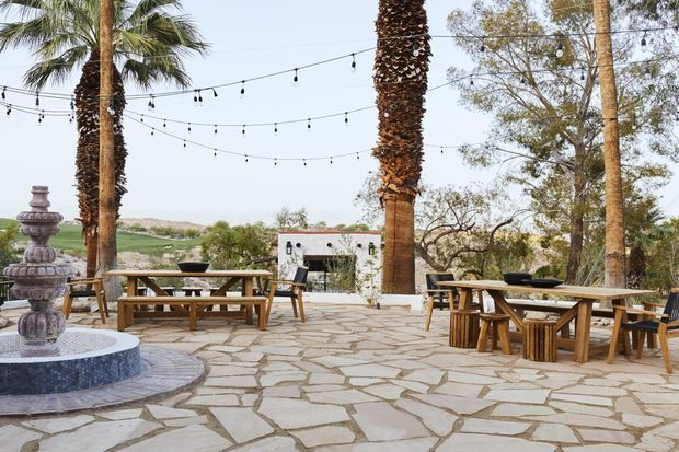 A spacious outdoor terrace or courtyard paved with irregular natural stone, furnished with rustic wooden picnic-style dining tables and benches. Tall palm trees and mature landscaping surround the area, with string lights overhead and a tiered stone fountain in the foreground. A low white building and open landscape are visible beyond the terrace.