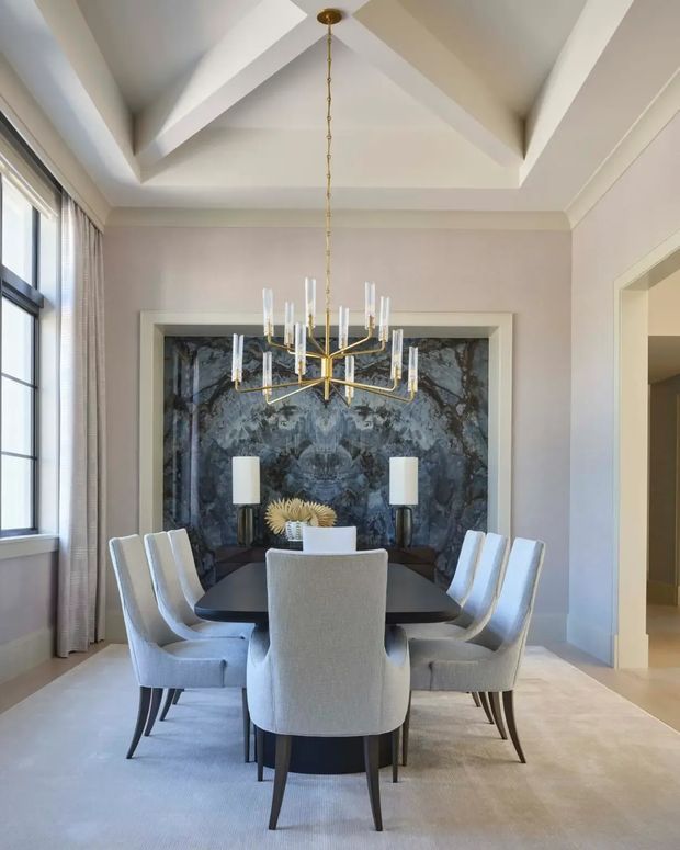 Elegant formal dining room with a dark dining table surrounded by upholstered dining chairs, a dramatic dark stone feature wall, sculptural brass chandelier, tall window, pale neutral walls, and a coffered or vaulted ceiling with strong geometric beams.