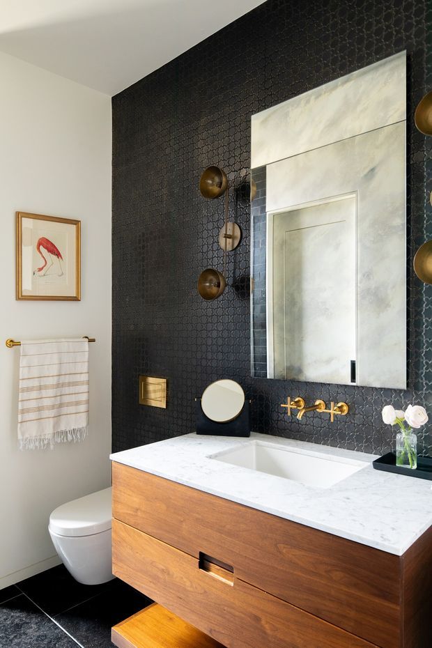 A compact, refined bathroom featuring a floating warm timber vanity with a white stone benchtop, integrated rectangular basin, and brass wall-mounted tapware. The main feature wall is clad in dark patterned tile or textured finish, complemented by a large pale framed mirror and globe-style wall sconces. A white toilet, striped hand towel, framed artwork, and black floor tiles complete the space.