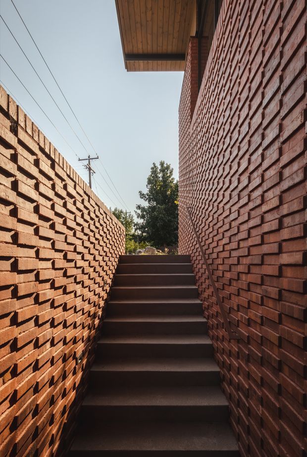 A narrow exterior stairway rises between two tall red brick walls with strongly textured projecting brickwork. Dark concrete steps lead upward toward a small landscaped opening, while a timber-lined roof overhang projects above the right wall. Utility lines and a tree are visible beyond the stair enclosure. The image emphasizes the passage-like architectural form, warm brick masonry, and dramatic vertical perspective.