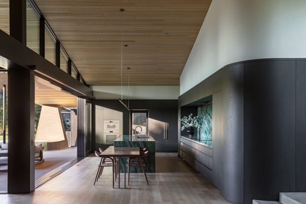 A refined open-plan kitchen and dining area with dark floor-to-ceiling cabinetry, a central island, timber dining table, and sculptural pendant lighting. Full-height glazing opens the room to an adjoining outdoor living area, while a warm timber-lined ceiling and pale timber flooring balance the dark joinery. The space has a clean contemporary architectural character with strong indoor-outdoor connection.
