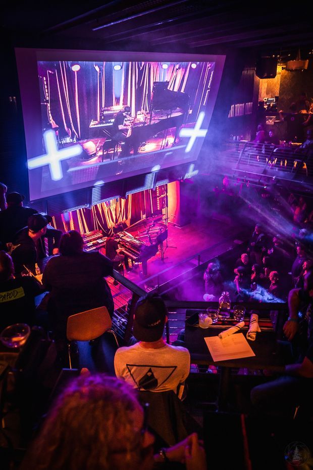 A dark, atmospheric live music and entertainment venue viewed from elevated seating. A brightly lit stage with performers and instruments is framed by a large projection screen, while tables, railings, and a seated audience fill the foreground and surrounding levels. Purple, magenta, blue, and warm orange stage lighting creates a dramatic nightclub or concert ambience.