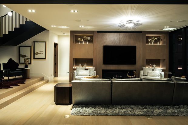 A sophisticated contemporary living room viewed from behind a large dark sectional sofa. The room features a built-in timber media wall with recessed illuminated display niches, a wall-mounted television, a linear fireplace, two upholstered lounge chairs, and layered ambient lighting. Pale timber flooring, dark joinery, a textured grey rug, and a sculptural ceiling light create a refined, dramatic atmosphere. A stairway and small adjacent seating area are visible to the left.