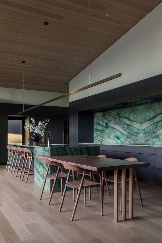 A sophisticated open-plan kitchen and dining area with a long timber dining table and coordinated wooden chairs in the foreground. The kitchen features dark timber cabinetry, a large green-veined stone island and matching dramatic stone splashback, with a timber-lined ceiling, slim linear pendant lighting, and pale timber flooring. The space has a dark, refined palette balanced by natural wood and richly patterned stone.