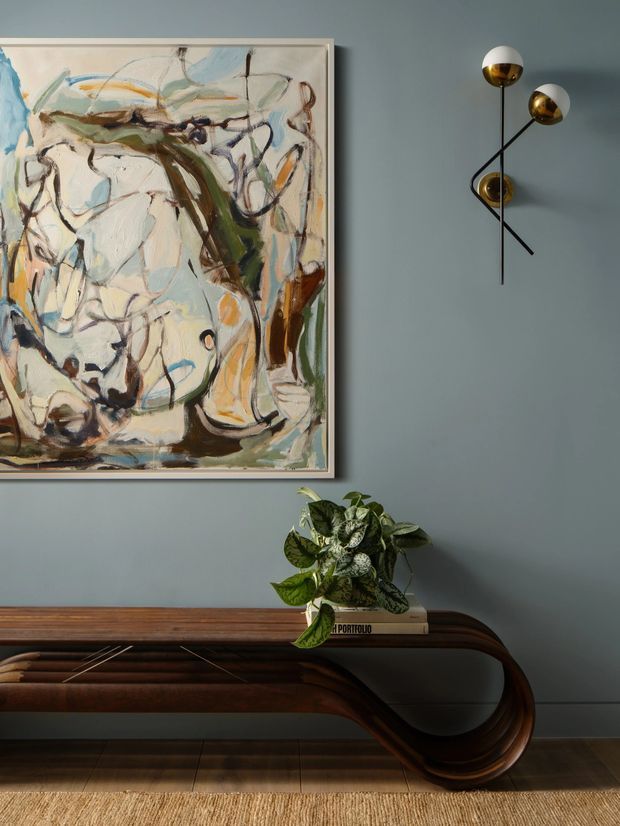 A stylish interior vignette featuring a dark timber mid-century console with sculptural curved legs, set against a muted blue-grey wall. Above it hangs a large abstract painting in a pale frame, accompanied by a sculptural black-and-brass wall light. A leafy potted plant and books sit on the console, with timber flooring and a woven rug below. The composition is warm, refined, and art-focused.