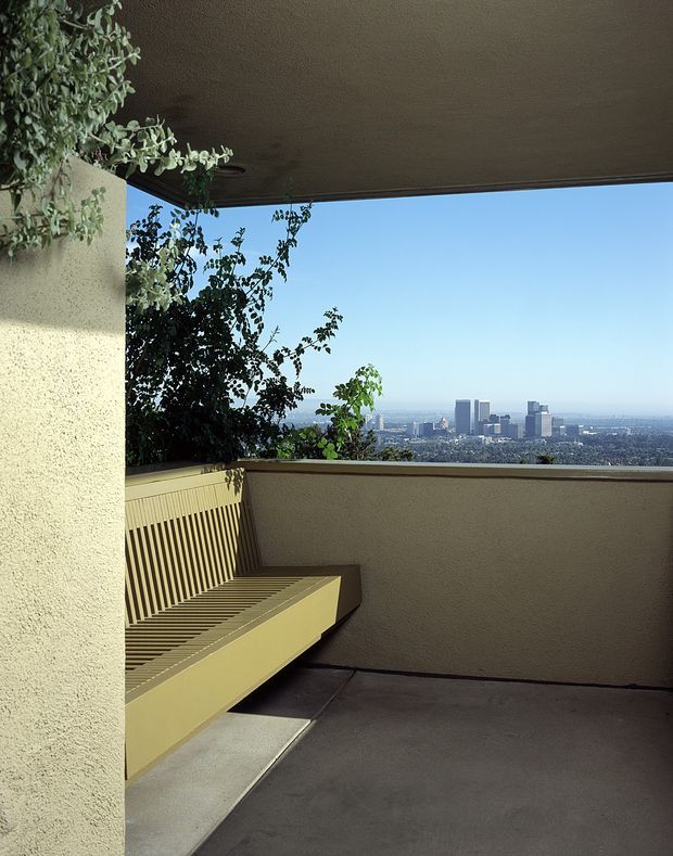 A covered concrete balcony or terrace with a built-in slatted bench, textured stucco walls, and a broad elevated view across a dense city skyline. Mature leafy planting partially frames the outlook, while the simple neutral finishes and strong horizontal roof and balustrade lines create a restrained modernist composition.