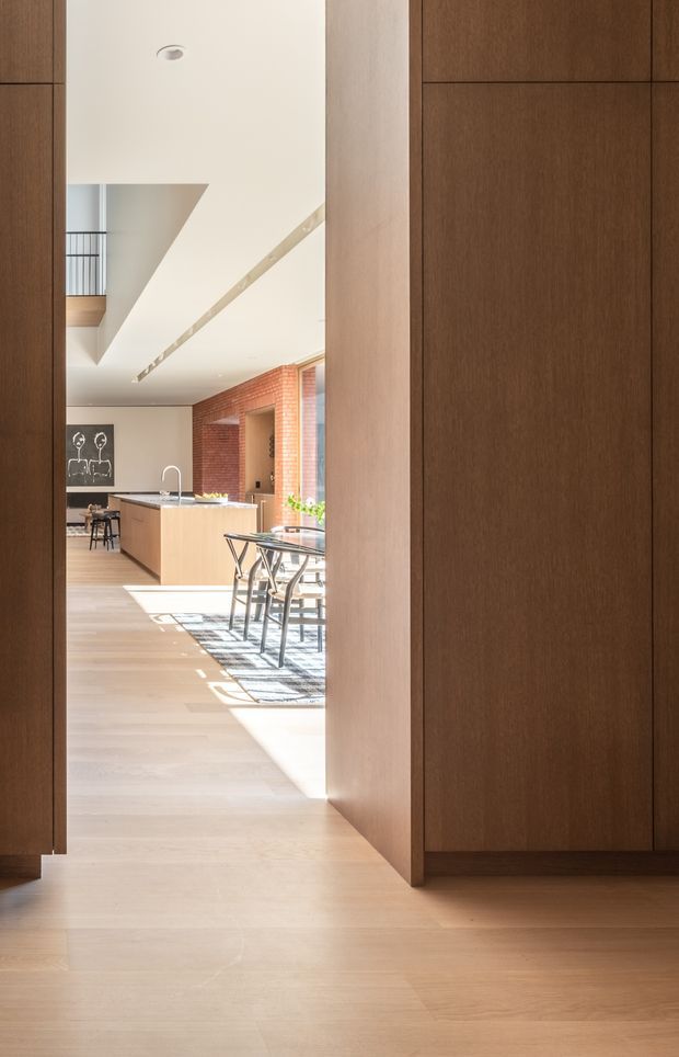A bright, contemporary open-plan kitchen and dining area viewed through a wide opening framed by dark timber built-in joinery. The space features pale timber flooring, light wood kitchen cabinetry with a dark benchtop, a dining table with black-framed chairs, a red brick feature wall, and abundant natural light. A linear recessed ceiling light and a partial stair or mezzanine opening add architectural interest.