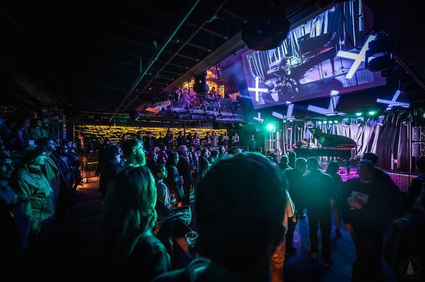 A dark, crowded indoor live-music venue or nightclub with a stage, grand piano, performers, colorful blue and green lighting, large projection screens, and an audience gathered across multiple levels. The space has an industrial entertainment-venue atmosphere with exposed ceiling services and theatrical lighting.