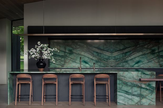 A sophisticated contemporary kitchen and entertaining space featuring a long island with four timber bar stools, dark vertically textured cabinetry, and a dramatic green-veined stone backsplash and waterfall island end. A black linear rangehood or feature band runs beneath pale upper cabinetry, while warm timber ceiling boards and shelving add contrast. Brass-toned taps and restrained styling create a refined, moody atmosphere.