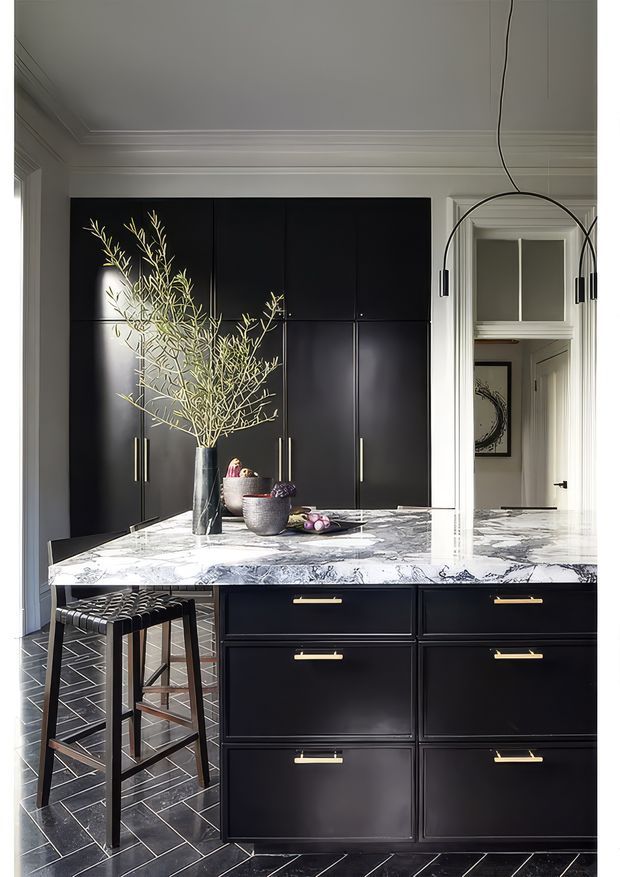 A sophisticated dark-toned kitchen centered on a large island with a dramatic white and grey veined stone benchtop. Floor-to-ceiling black cabinetry with slim brass handles provides extensive storage, while black bar stools sit at the island. A sculptural branch arrangement, bowls and fruit add decorative detail. The room features white traditional-style wall and ceiling mouldings, a geometric dark tiled floor, and a large arched black pendant light.