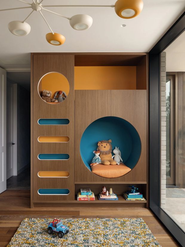A playful children's room feature wall with custom timber cabinetry and integrated display niches. The built-in joinery includes a large circular blue reading or toy alcove with cushions and soft toys, a smaller circular display opening, colourful rounded inset handles or shelves, and open book storage below. A graphic multi-arm ceiling light, timber flooring, and a multicoloured rug complete the warm, colourful interior. Full-height glazing and dark framing are visible to the right.