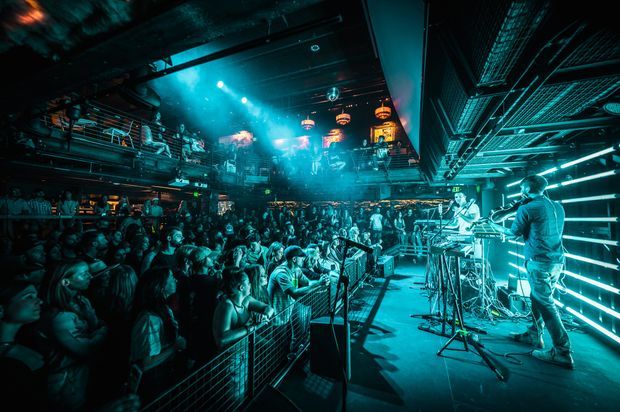 A dark, high-energy indoor concert or nightclub venue packed with a large crowd. The camera faces a brightly lit stage where musicians perform, with dramatic cyan-blue lighting, haze, exposed overhead services, metal railings, an upper-level balcony, and industrial metal grilles and structures throughout the space.