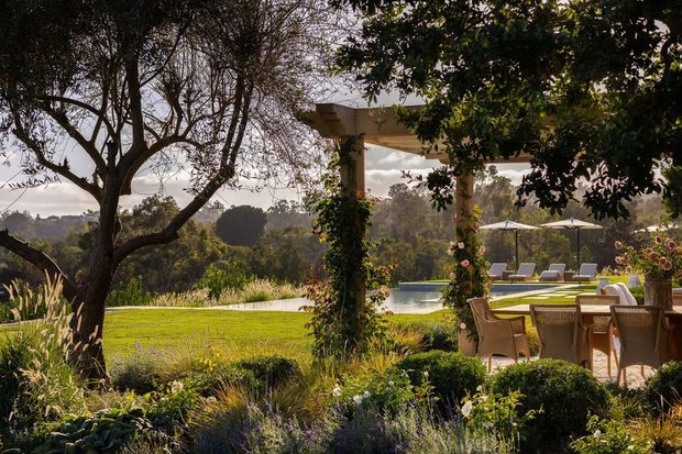 A landscaped outdoor entertaining area with a timber dining setting beneath a vine-covered pergola, overlooking a lawn, swimming pool, and wooded landscape. Lush ornamental planting and mature trees create a relaxed garden setting.
