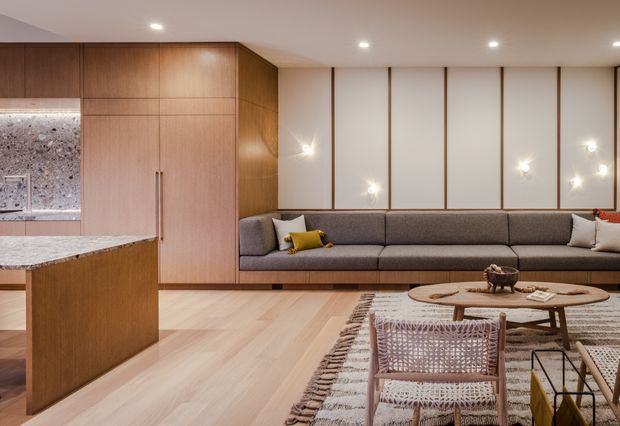 Warm, open-plan contemporary living area with an integrated kitchen. The room features extensive light timber cabinetry and flooring, a long built-in grey upholstered sofa, white wall panels framed with timber battens, decorative wall sconces, and layered round coffee tables over a textured rug. A stone kitchen island and stone backsplash are visible on the left.