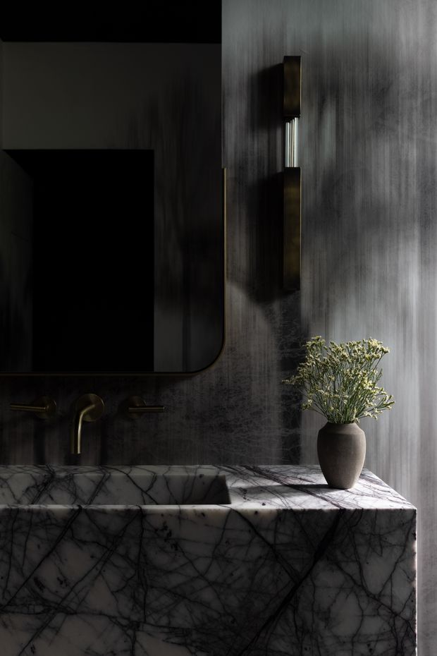 A dramatic, moody bathroom vanity vignette featuring a dark marble or stone wall and a substantial white marble vanity with bold dark veining. A wall-mounted bronze or dark metal faucet sits beneath a large rounded-corner mirror, beside a slim vertical wall light. A sculptural dark vase with delicate foliage adds a natural accent. The space has a luxurious, high-contrast contemporary aesthetic.
