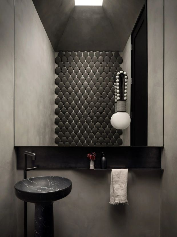 A compact, moody bathroom with dark grey plastered walls and a dramatic black stone pedestal basin. The vanity wall features a black backsplash and floating shelf, while the rear wall is covered in rounded dark tile arranged in a scalloped pattern. A large black-framed mirror, sculptural pendant light, black tapware, and a skylight complete the refined monochromatic interior.