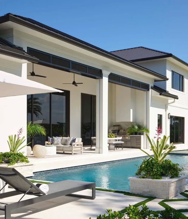 Bright exterior view of a contemporary two-storey white-rendered home with dark tiled hipped roofing, large glazed openings, covered outdoor entertaining areas, ceiling fans, an outdoor kitchen, and a swimming pool with landscaped raised planters and poolside loungers.
