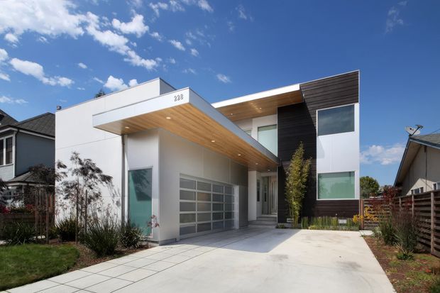 Modern two-storey residential house photographed from the driveway, featuring a clean white rendered facade, dark horizontal cladding, a broad flat roof and projecting timber-lined soffit over the garage and entry. Large glazed openings, a contemporary garage door, paved driveway and landscaped front garden are visible, with neighbouring homes on either side.