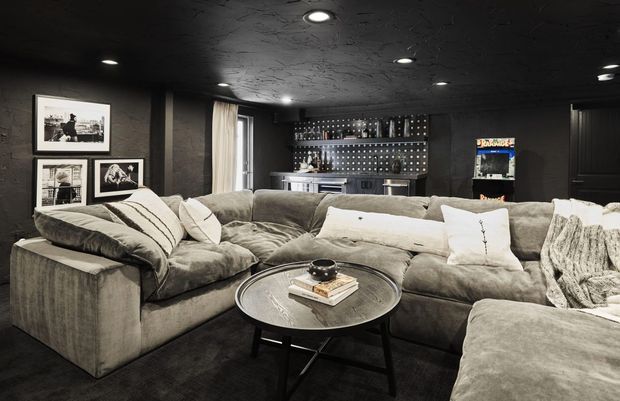 A dark, moody home media room with a large plush sectional sofa, round coffee table, framed wall art, recessed ceiling lights, and a built-in bar or display wall at the rear. The space features charcoal textured walls and ceiling, soft grey upholstery, and a pinball machine, creating a relaxed entertainment-focused atmosphere.