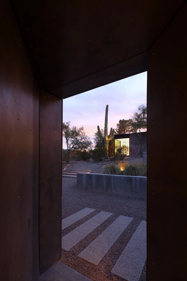 A dark, narrow architectural passage or entry framed by deep brown metal or timber walls and ceiling, opening toward a landscaped courtyard at dusk. The view includes a gravel forecourt with broad rectangular stepping slabs, a concrete planter wall, desert grasses, and a tall sculptural saguaro cactus. A low modern building with illuminated glazing is visible beyond, creating a dramatic threshold between the enclosed entry and the desert garden.